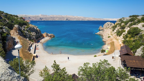 A coastal beach with clear blue waters, rocky terrain, and a few buildings including a small structure with a red roof.
