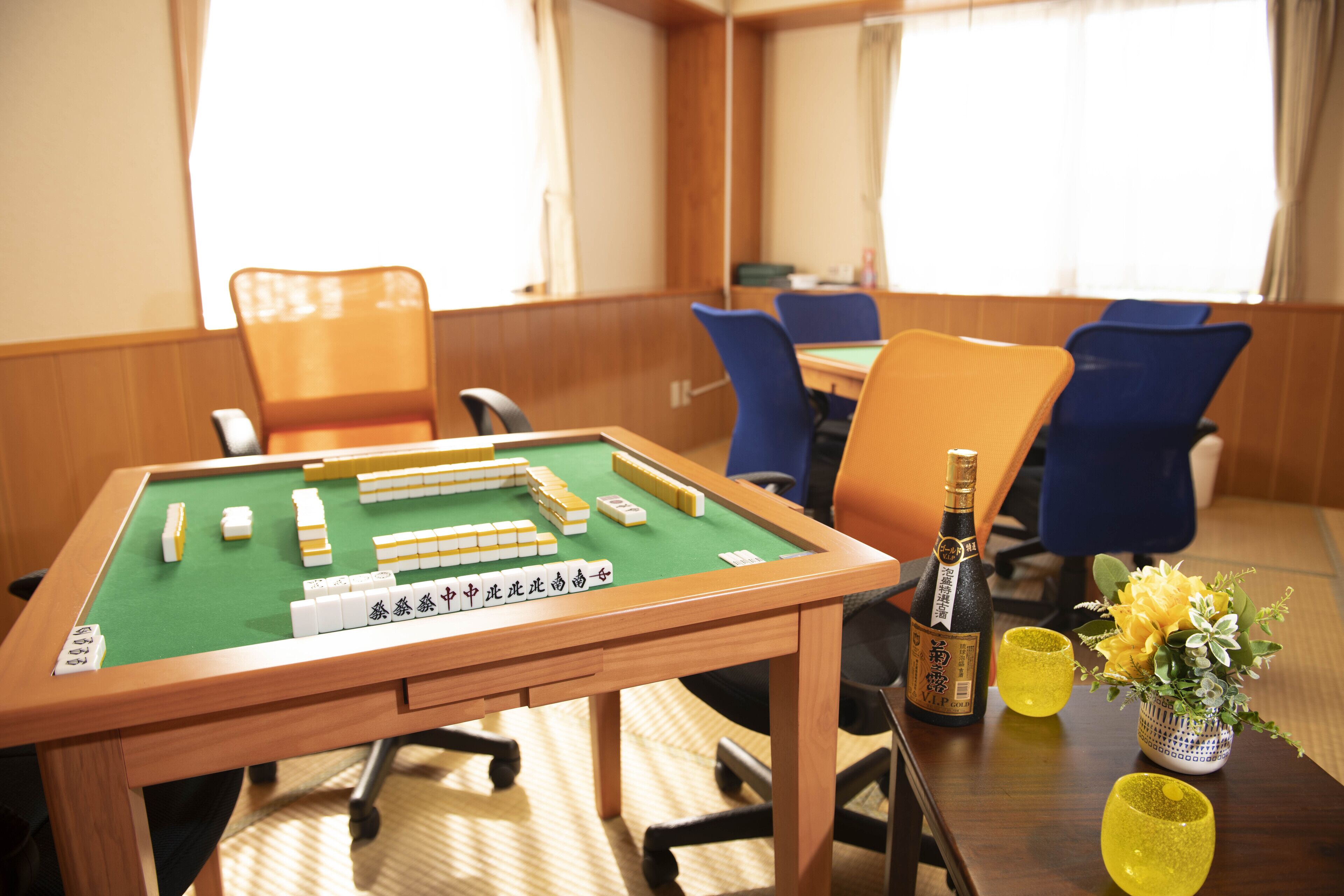 Een mahjongtafel met tegels, een fles en twee glazen op een houten tafel in een kamer met blauwe stoelen en een raam.