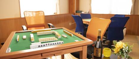 Een mahjongtafel met tegels, een fles en twee glazen op een houten tafel in een kamer met blauwe stoelen en een raam.