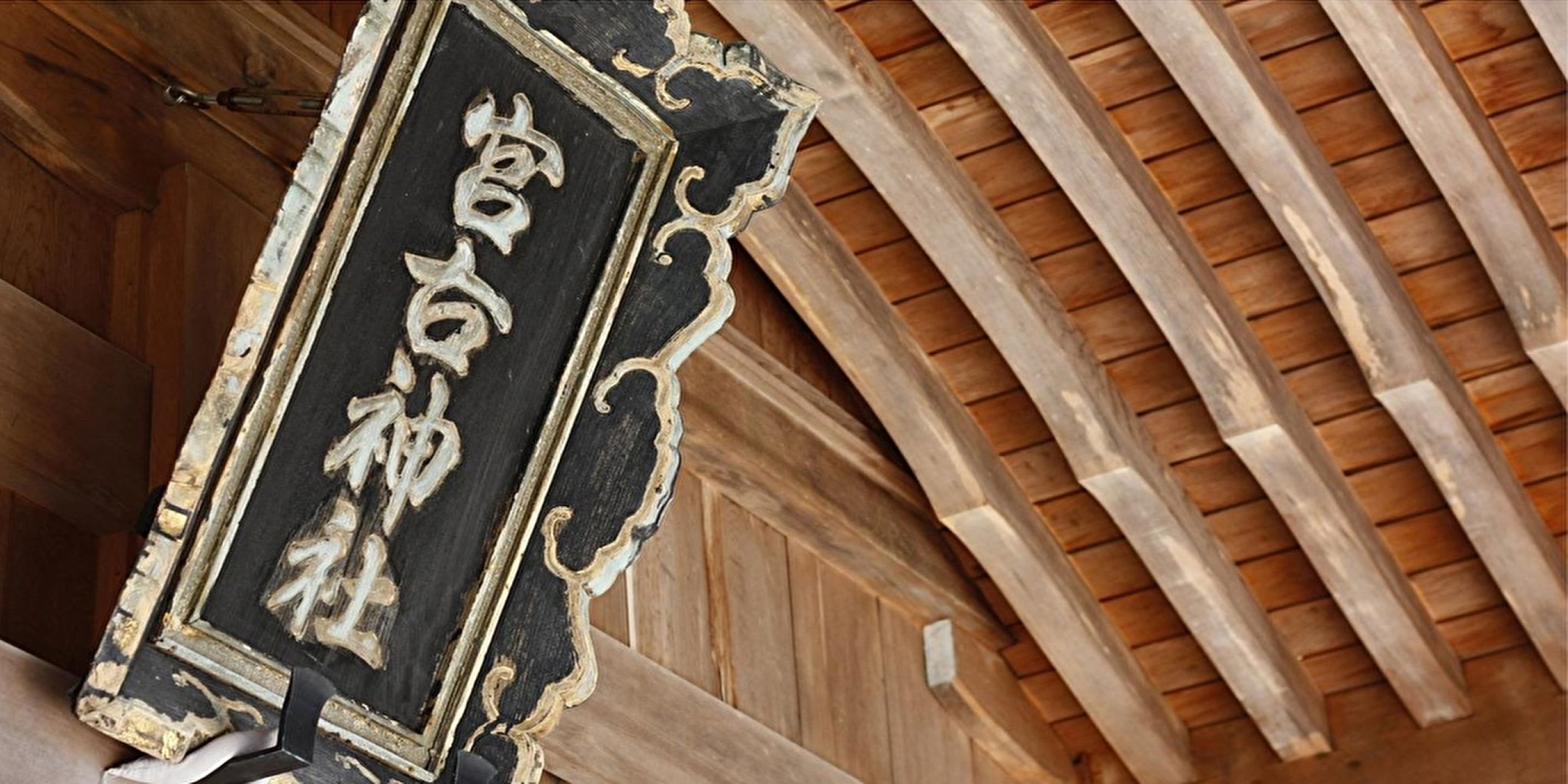 Ein schwarzes und goldenes, verziertes Schild mit japanischen Schriftzeichen, das an einem Holzbalken befestigt ist.