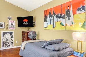 A bedroom with a bed, a TV mounted on the wall, a fan, and artwork. - Charming 2 bedroom cottage with private pool in the heart of the Texas Hill Coun (Johnson City)