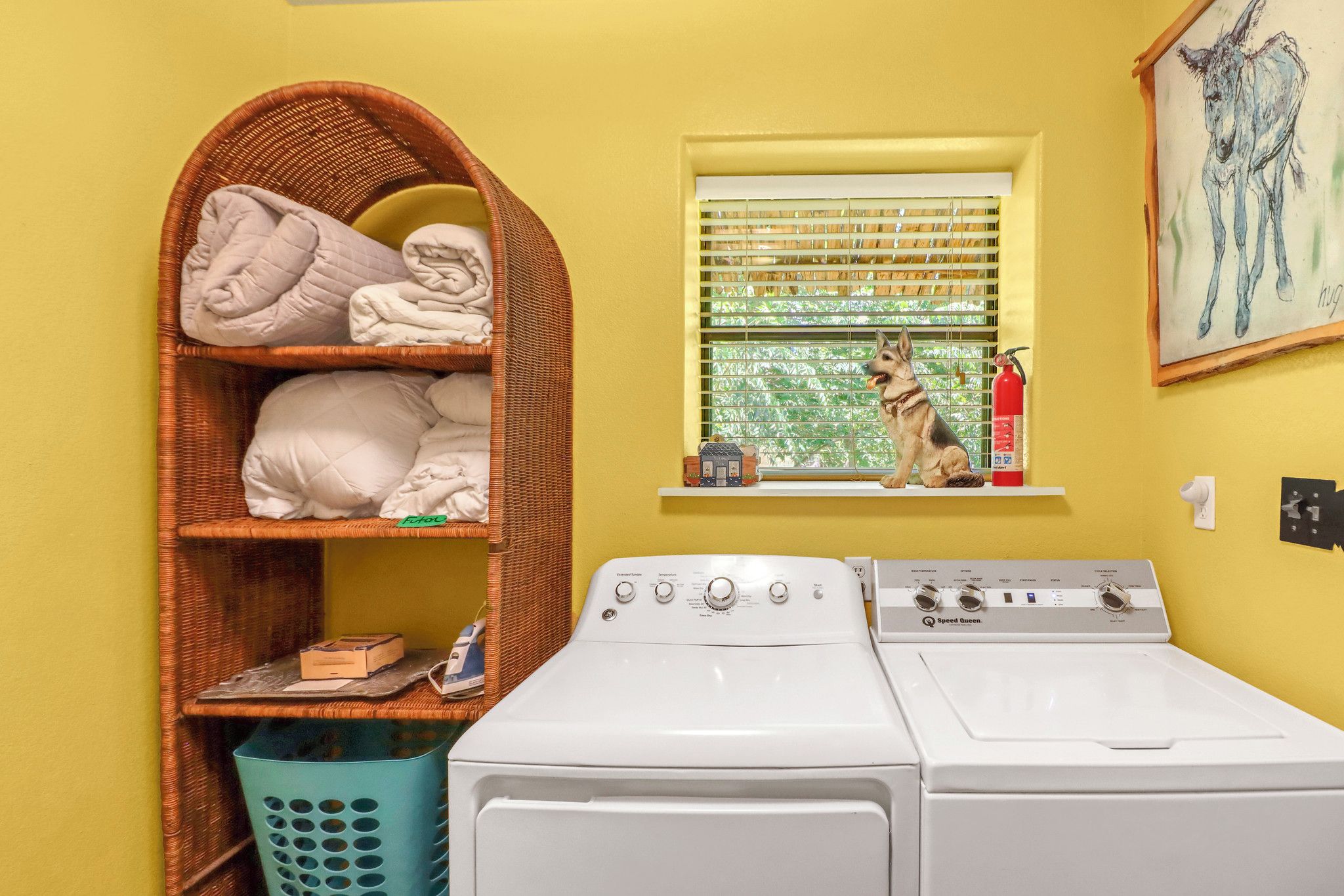 A laundry area with a washing machine, a wicker shelf with folded towels, a window with blinds, and a fire extinguisher.
