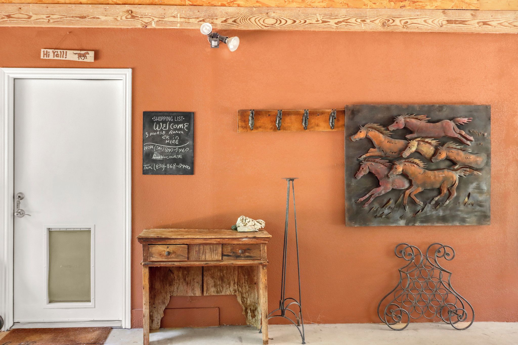 A room with a wooden desk, a chalkboard with a shopping list, a horse-themed painting, and a metal sculpture.