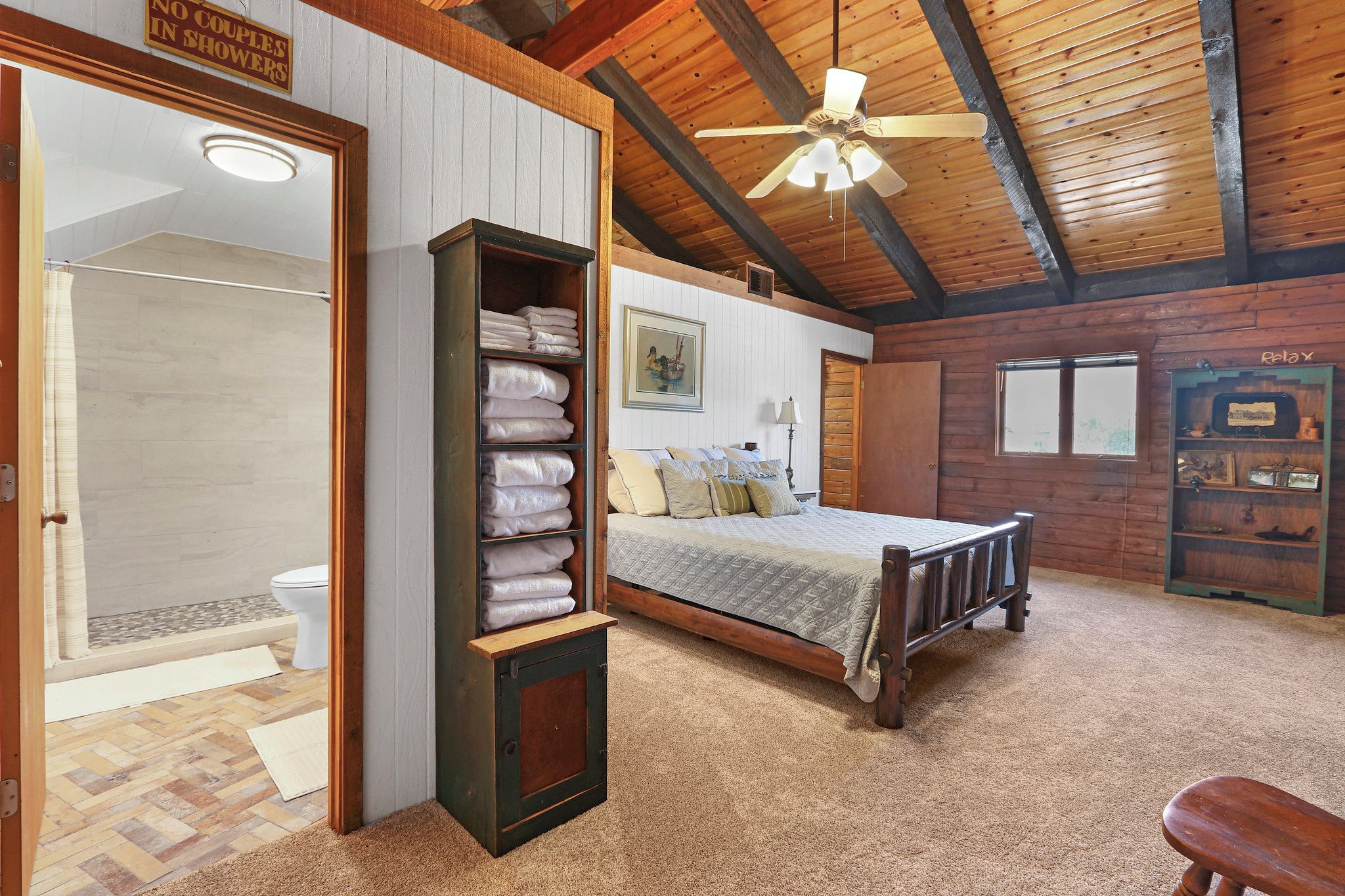 Un dormitorio con techo de madera, una cama, un estante con toallas dobladas, una ducha y un letrero que indica que no se permiten parejas en la ducha.