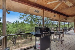 An outdoor deck with a barbecue grill, seating area, and a view of trees and hills. - Spacious 2/2 - ON the Cypress Creek with private hot tub! (Wimberley)