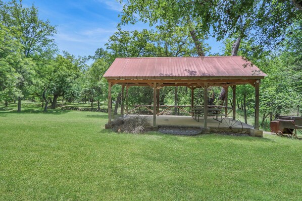 A gazebo with a metal roof and wooden frame, surrounded by a grassy area and trees.