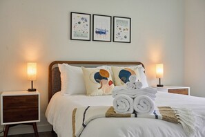 Una cama bien hecha con sábanas blancas, dos almohadas con diseños abstractos y una pila de toallas enrolladas. Hay dos mesitas de noche con lámparas, y tres obras de arte enmarcadas en la pared.