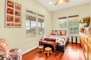 Un dormitorio con cama, dos ventanas, ventilador de techo y un taburete pequeño.