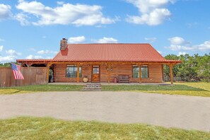 A log cabin with a red-tiled roof, a front porch with a bench, and an American flag. - 3 bedroom pet friendly log cabin with private hot tub! (Wimberley)