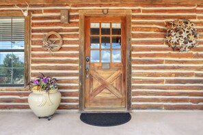 A wooden log cabin with a large wooden door, a window with blinds, and a potted plant with flowers.