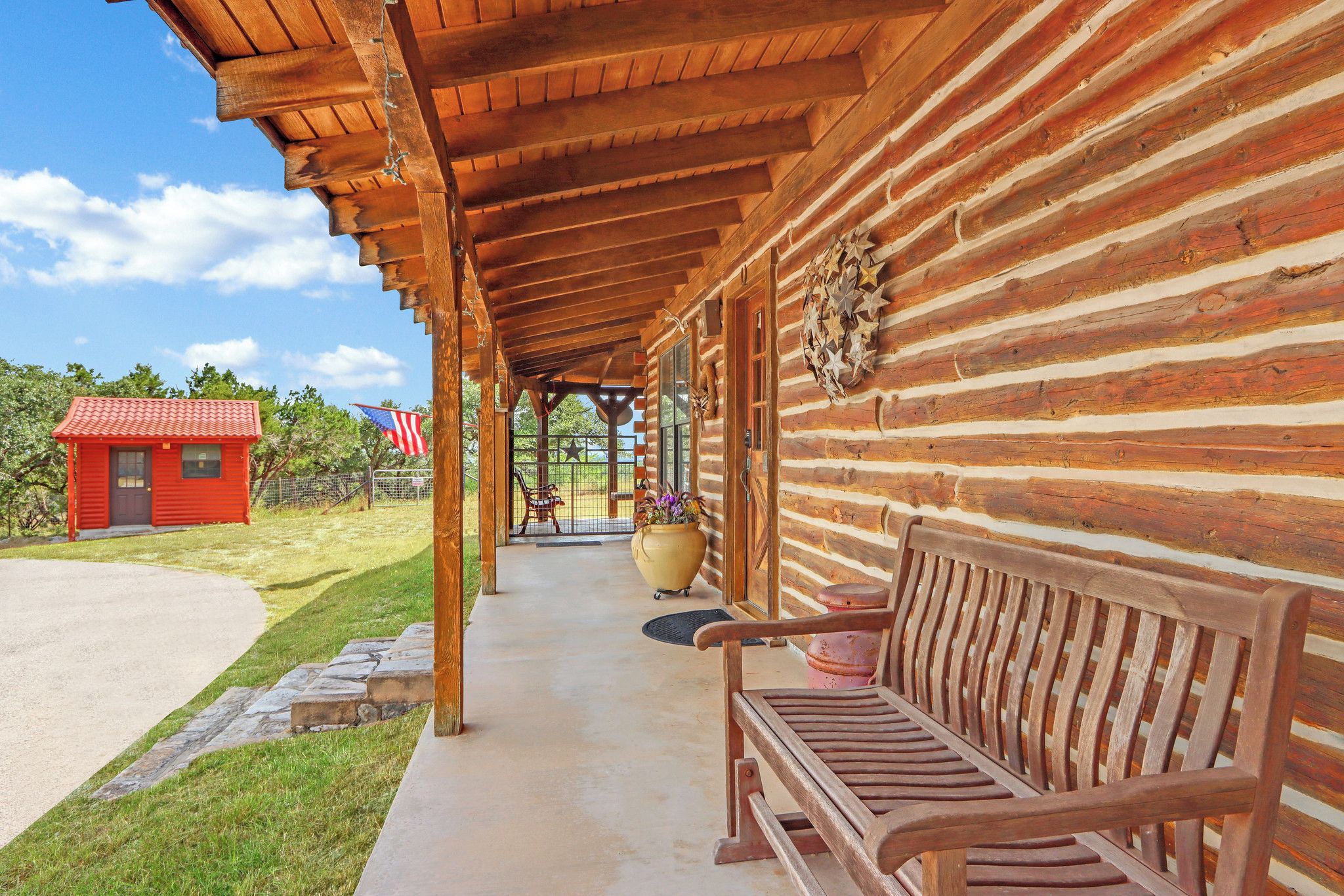 A wooden log cabin with a covered porch, a red building in the background, and a bench.