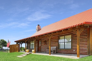 A log cabin with a red-tiled roof, large windows, and a porch with benches. - 3 bedroom pet friendly log cabin with private hot tub! (Wimberley)