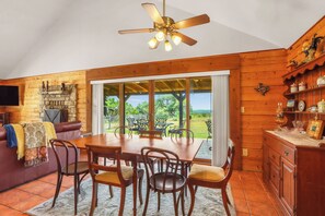 A wooden interior with a dining table, chairs, a fireplace, and a large window overlooking a garden.