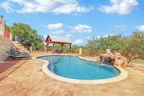Una piscina con una cascada, una zona de estar cubierta y un muro de piedra.