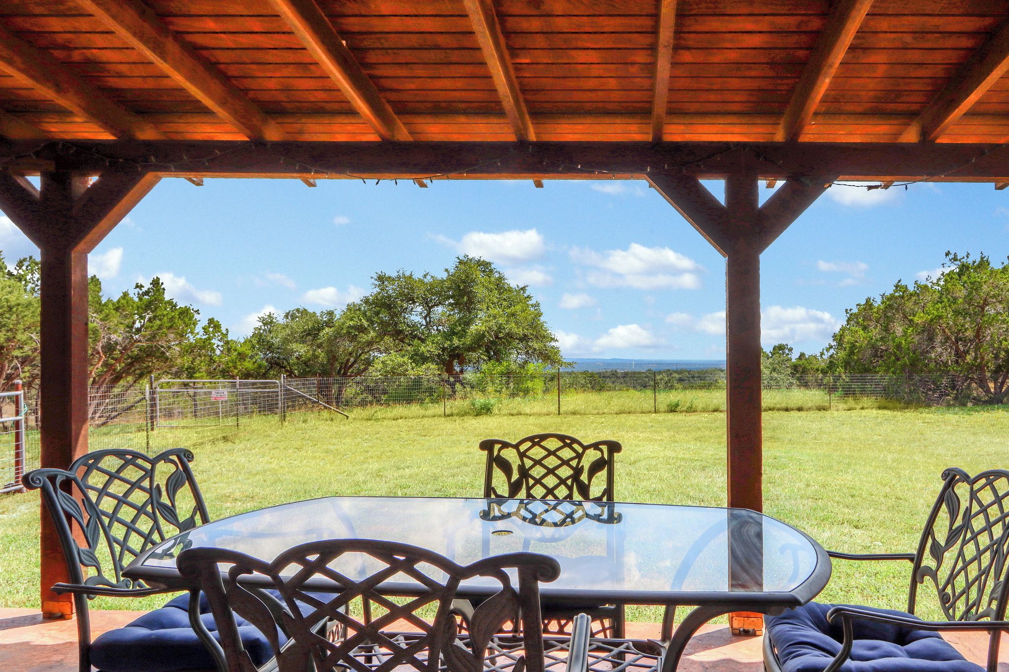 Un espacio comedor exterior cubierto con una mesa de vidrio y sillas de metal, con vistas a un paisaje con césped, árboles y una valla.