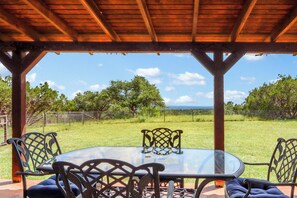 A covered outdoor dining area with a glass table and metal chairs, overlooking a grassy landscape with trees and a fence. - 3 bedroom pet friendly log cabin with private hot tub! (Wimberley)