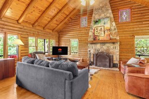 Interior de una cabaña de troncos con chimenea de piedra, televisor y sofá.