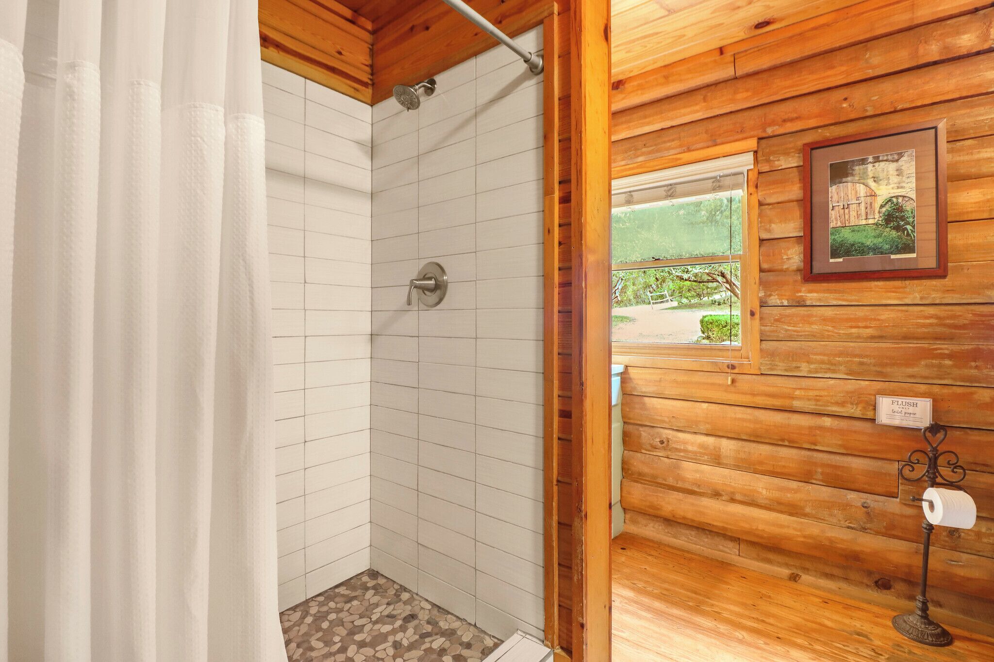 Ein Badezimmer einer Holzhütte mit Dusche, Toilettenpapierhalter und einem Fenster mit Blick auf Grünflächen.