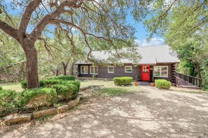 A rustic log cabin with a red door, surrounded by a well-maintained garden and a large tree.