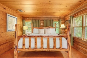 A wooden cabin bedroom with a bed, two bedside lamps, and a window with blinds. - Popular 2BR Log Home on the Creek with a Hot Tub! (Wimberley)