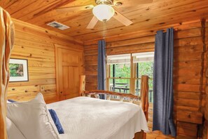A wooden cabin bedroom with a bed, a ceiling fan, a window with curtains, and a framed picture on the wall.
