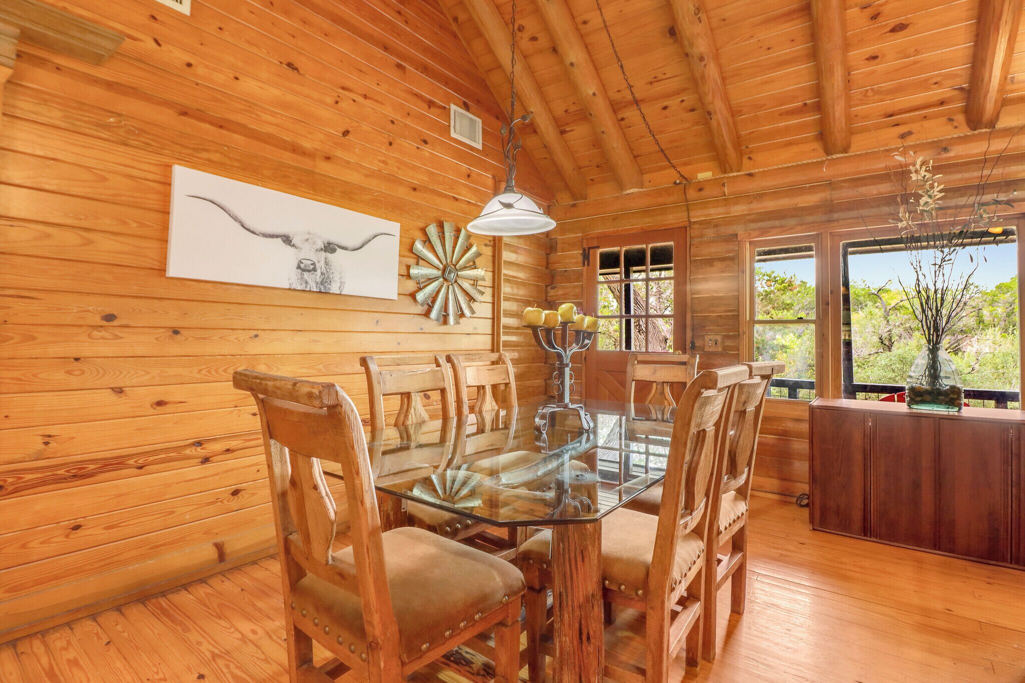 A wooden interior with a dining table, chairs, a chandelier, and a window with a view.