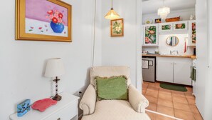 Una sala de estar con sofá, lámpara, un cuadro enmarcado y una cocina visible a través de un vano abierto.