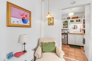 Una sala de estar con sofá, lámpara, un cuadro enmarcado y una cocina visible a través de un vano abierto.