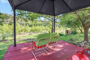 A covered outdoor seating area with two chairs and a table, surrounded by greenery and trees.
