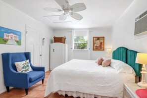 A bedroom with a bed, a blue armchair, a white ceiling fan, a window with a view, and a painting on the wall. - Darling cottage on the Blanco River. Minutes from downtown Wimberley! (Wimberley)