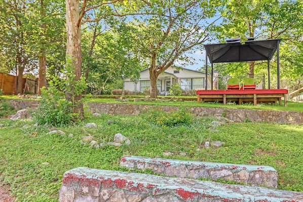 Patio trasero con zona de estar cubierta, un muro de piedra y una casa al fondo.