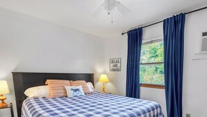 Un dormitorio con cama, mesita de noche, lámpara, ventana con cortinas y un cartel en la pared.