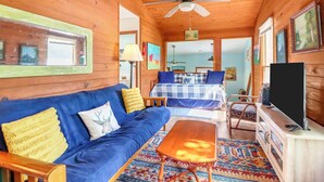 Interior acogedor de una cabaña con techo de madera, un sofá, una cama con colcha azul y blanca a cuadros, un televisor y una alfombra estampada.