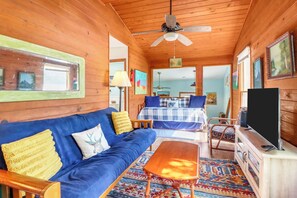 Interior acogedor de una cabaña con techo de madera, un sofá, una cama con colcha azul y blanca a cuadros, un televisor y una alfombra estampada.