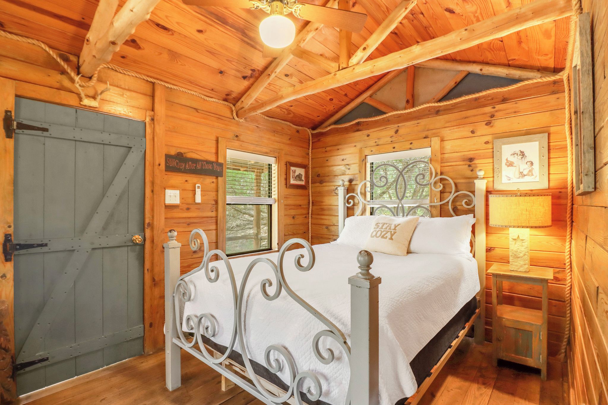 A wooden cabin bedroom with a bed, a window, a door, and a small bedside table with a lamp.