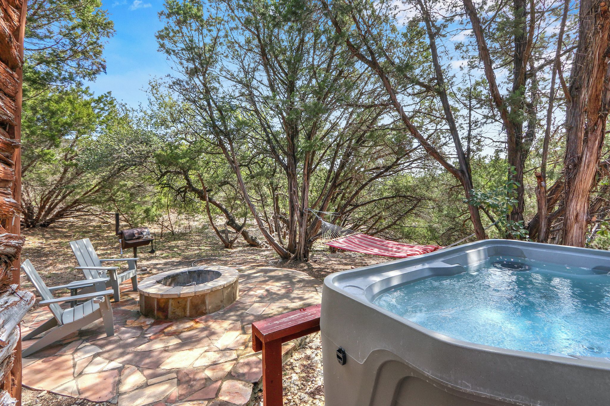 An outdoor hot tub area with stone flooring, a fire pit, and two Adirondack chairs.