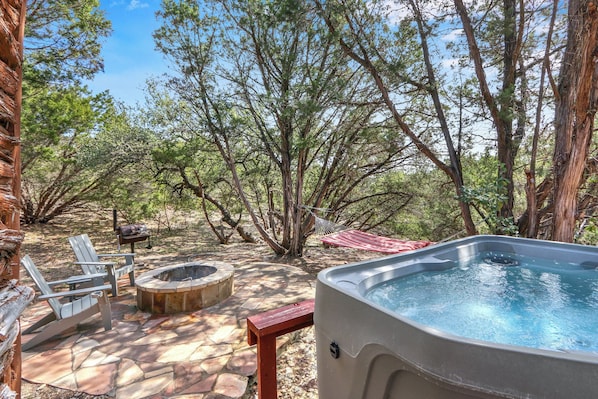 An outdoor hot tub area with stone flooring, a fire pit, and two Adirondack chairs.