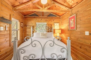 A wooden cabin bedroom with a metal bed frame, a window with a metal grill, and a wall-mounted light fixture. - Private, cozy and romantic cabin - a Wimberley favorite! Private hot tub! (Wimberley)