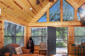 L’intérieur d’un chalet en bois, avec une grande porte ouverte donnant sur une terrasse extérieure, une bibliothèque, un fauteuil et une table offrant une vue sur les arbres à travers les fenêtres.