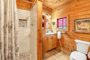 A wooden cabin bathroom with a shower, toilet, sink, and a small window with a pink curtain. - Private, cozy and romantic cabin - a Wimberley favorite! Private hot tub! (Wimberley)