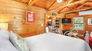Interior d'una cabana de fusta amb un llit, una televisió i una cuineta.