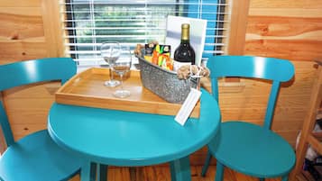 Uma mesa azul-turquesa com duas cadeiras combinando, uma bandeja de madeira com dois copos de vinho, uma garrafa de vinho e uma cesta com petiscos e um cartão.