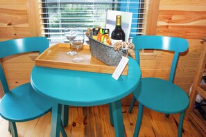 Une table de couleur turquoise, accompagnée de deux chaises assorties, un plateau en bois avec deux verres à vin, une bouteille de vin et un panier contenant des en-cas et une carte.