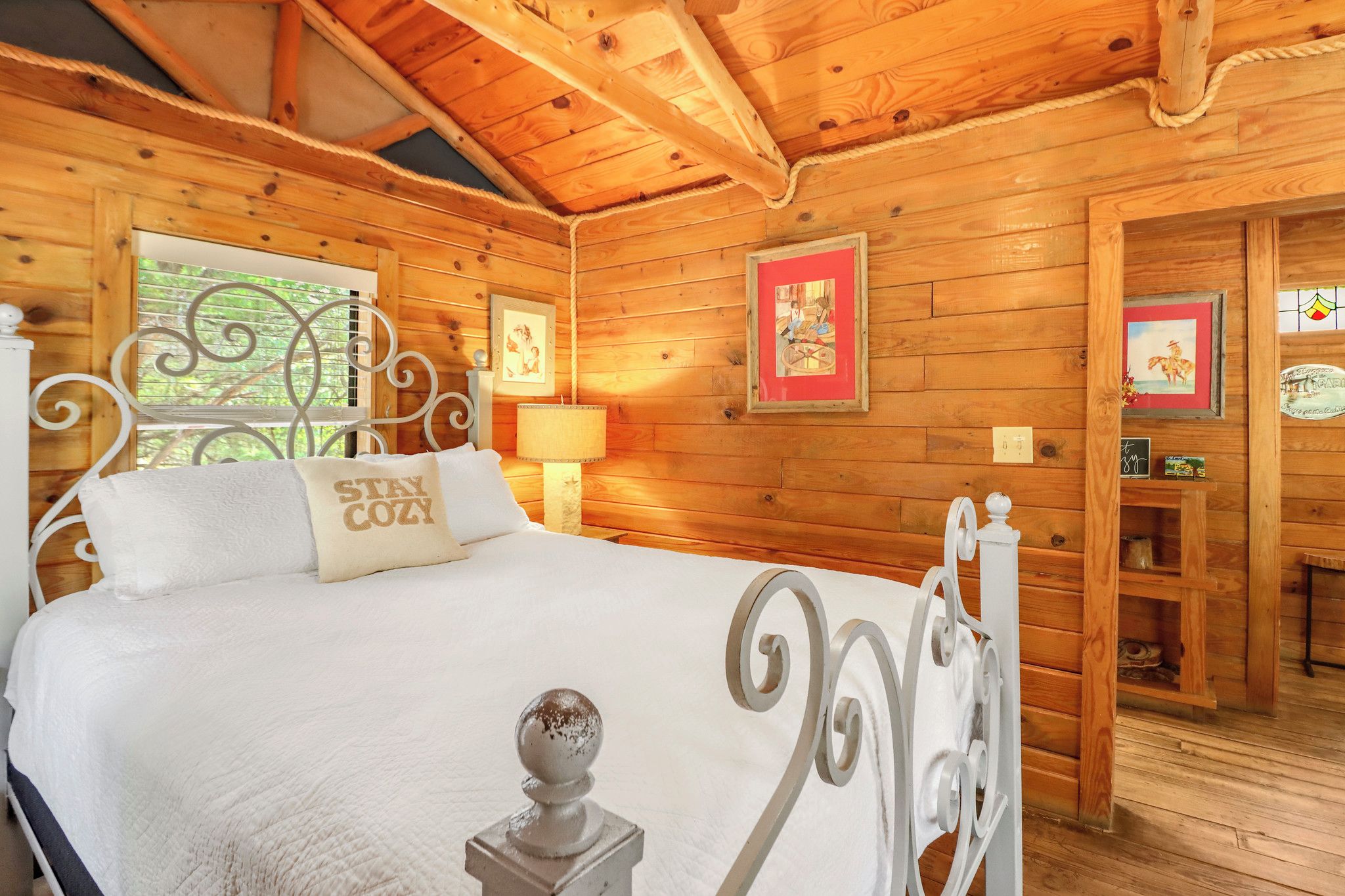 Una habitación de cabaña de madera con una cama, cabecera de metal, ventana con celosía metálica y pared con cuadros.