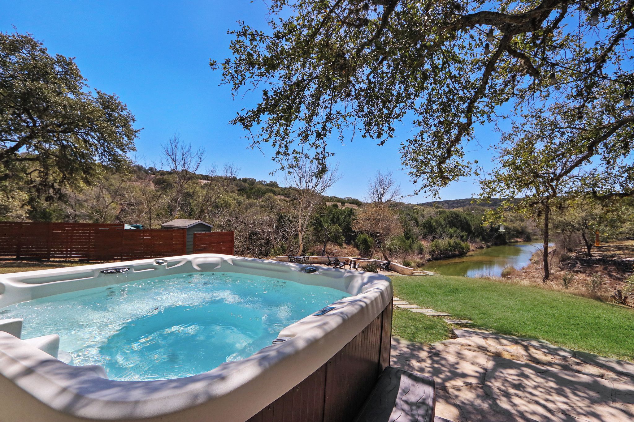 Un jacuzzi en un patio trasero con vista al río, rodeado de árboles y una valla de madera.