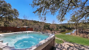 Un jacuzzi en un patio trasero con vista al río, rodeado de árboles y una valla de madera.
