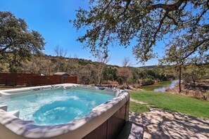 Un jacuzzi en un patio trasero con vista al río, rodeado de árboles y una valla de madera.