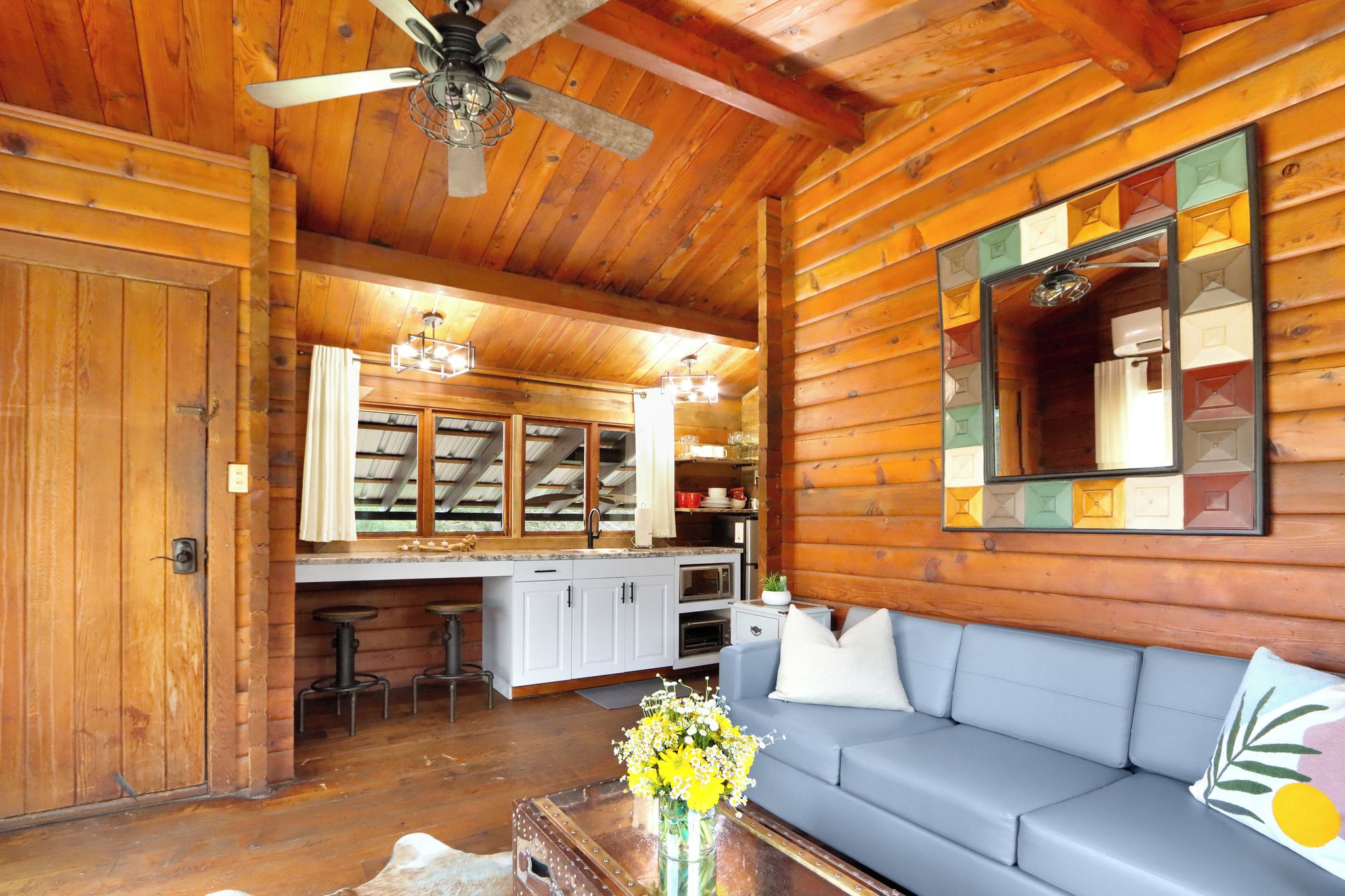 A log cabin interior with a wooden ceiling, a ceiling fan, a kitchenette, a sofa, and a mirror with a geometric frame.