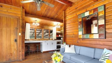 A log cabin interior with a wooden ceiling, a ceiling fan, a kitchenette, a sofa, and a mirror with a geometric frame.