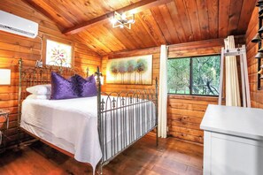 A wooden cabin bedroom with a bed, a nightstand, a window with a view of trees, and a ceiling light fixture. - Canyon Vista I Romantic, hot tub, log cabin (Wimberley)